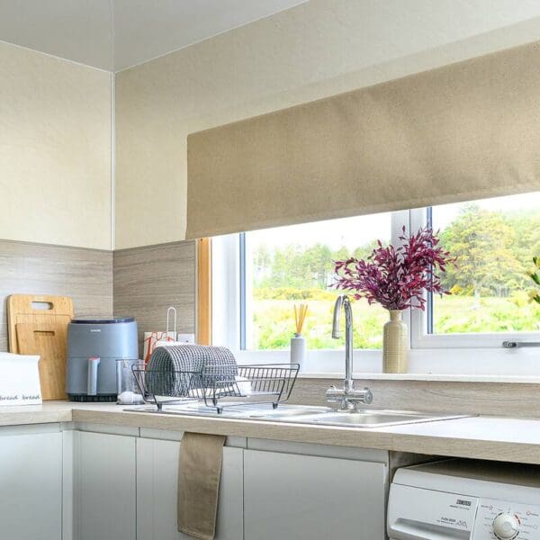Kitchen with white gloss cabinets, large window above sink with cream roller blind and purple flower display on windowsill