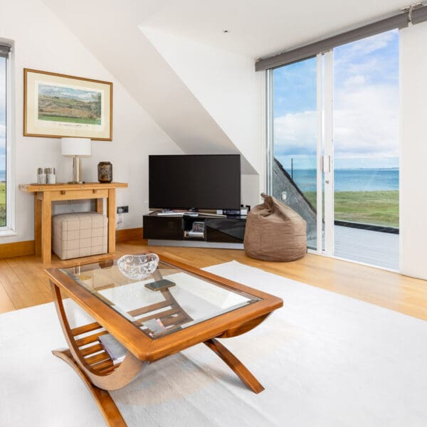 Living room with white walls and white rug on oak wood floor, white couch with tartan cushions, two picture windows looking out over elevated view of golf course and sea