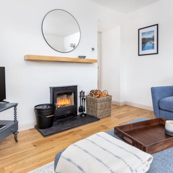 Living room with white walls, oak floor, blue material chair and foot stool, wood burning stove with fire lit, flatscreen tv on grey table