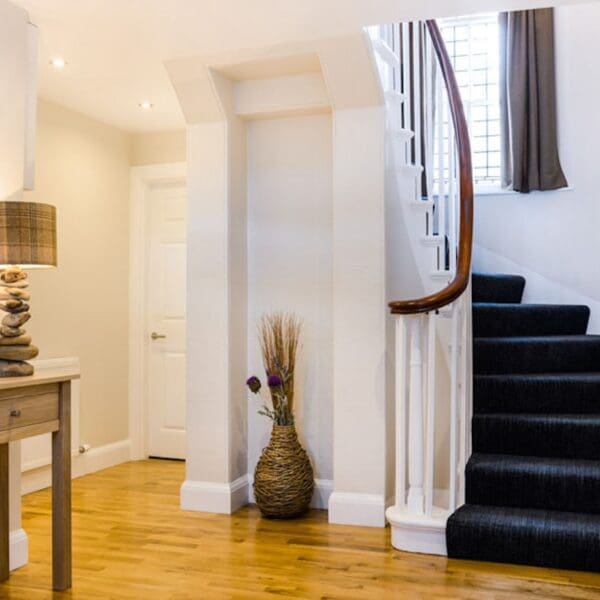 Hallway with lit lamp on wooden side table and spiral staircase with blue carpet, Burnside Cottage Dornoch