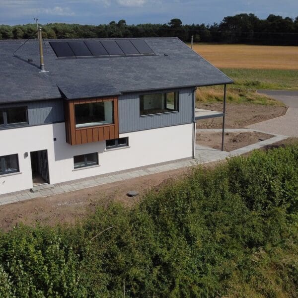 Exterior of modern build house with white, grey and wood cladding and grey roof with solar panels, surrounded by arable fields and woodland