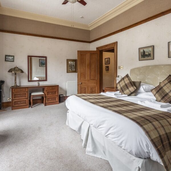 Bedroom with cream walls and carpet, pine furniture and brown tartan rug and cushions on bed