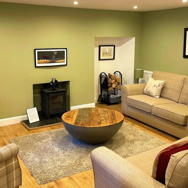 Living room in Carnegie Cottage, Dornoch with green painted walls, grey and light brown three piece suite, woodburning stove and wide screen tv