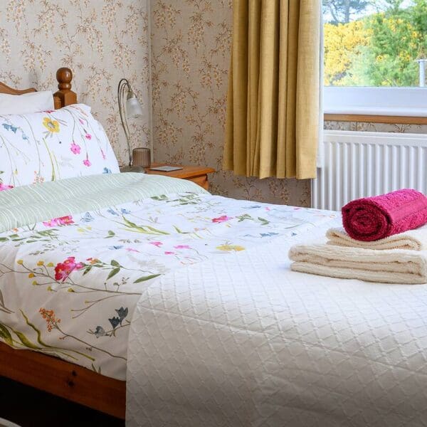 single bedroom with pine bed and floral duvet. Yellow curtains and window with view of yellow gorse.