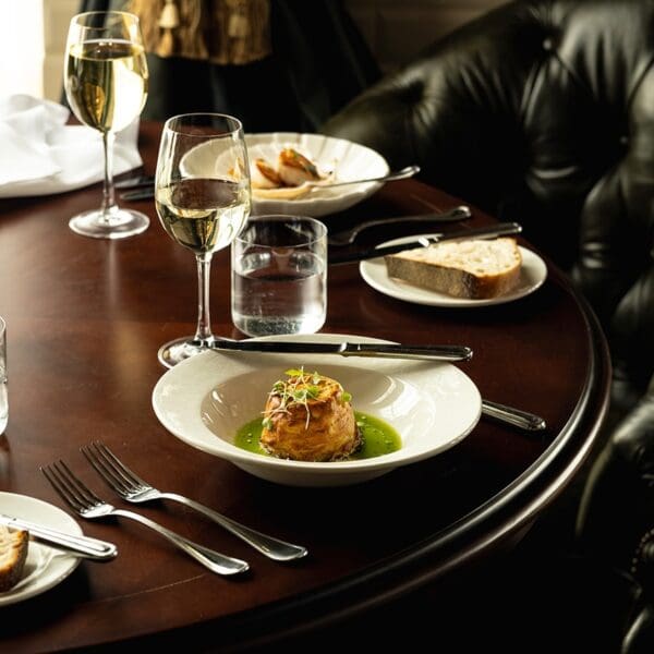 Plate of food in small white bowl set on dark wood table with bread on side plates and glasses of red and white wine. Green leather button back chair