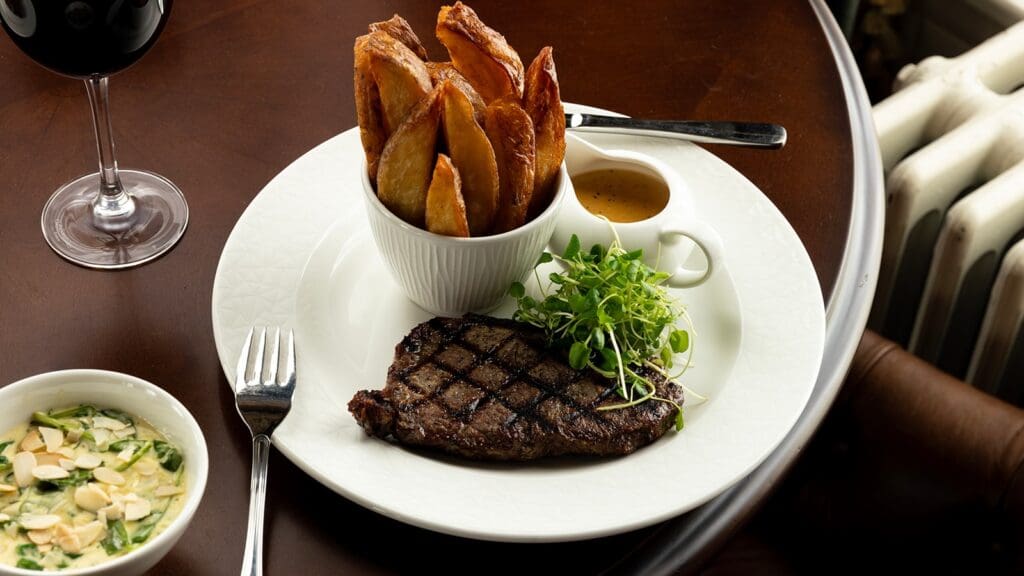 Seared stake on white plate with green salad, small jug of sauce and bowl of thick cut chips, set on dark wood tabletop alongside glass of red wine