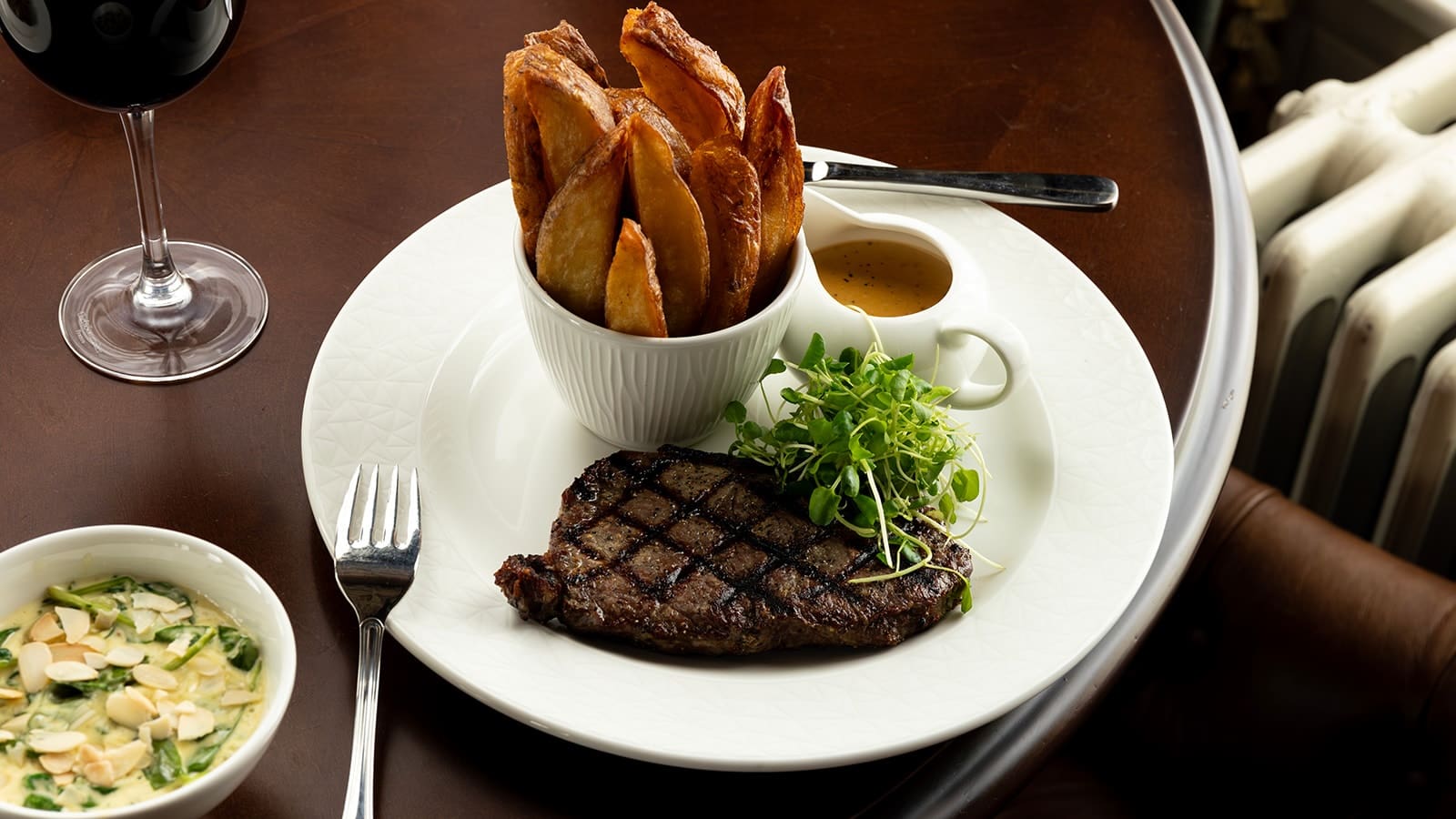 Seared stake on white plate with green salad, small jug of sauce and bowl of thick cut chips, set on dark wood tabletop alongside glass of red wine