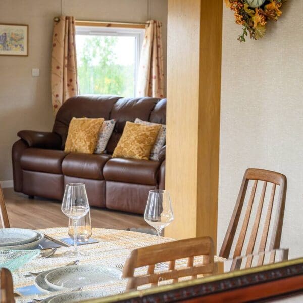 Image reflected in mirror of sitting room with dark brown leather couch and yellow cushions. In the foreground, dining room area with wooden chairs set around table with yellow check tablecloth and pale blue dinner service