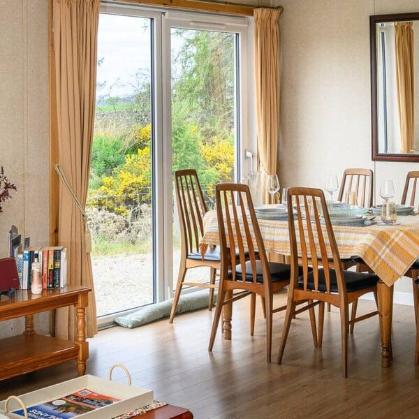 Dining room with pine table and chairs set with yellow check tablecloth and light blue dinner service. Patio doors, cream wall paper.