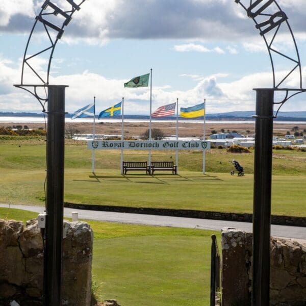 View through arched gateway towards sign for Royal Dornoch Golf club with five flags above, with Royal Dornoch Golf Club in lettering around arch.