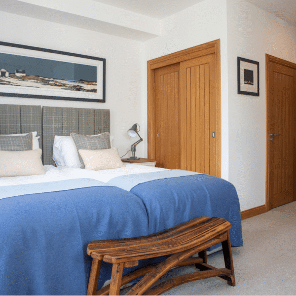 White twin bedroom with white sheets and blue blanket throws, pine stool at foot of bed and two pine doorways