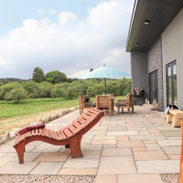 Exterior of grey clad modern bungalow, with paved patio area with wooden table and chairs and light blue parasol, overlooking grass field and woodland. Wooden recliner chair in foreground