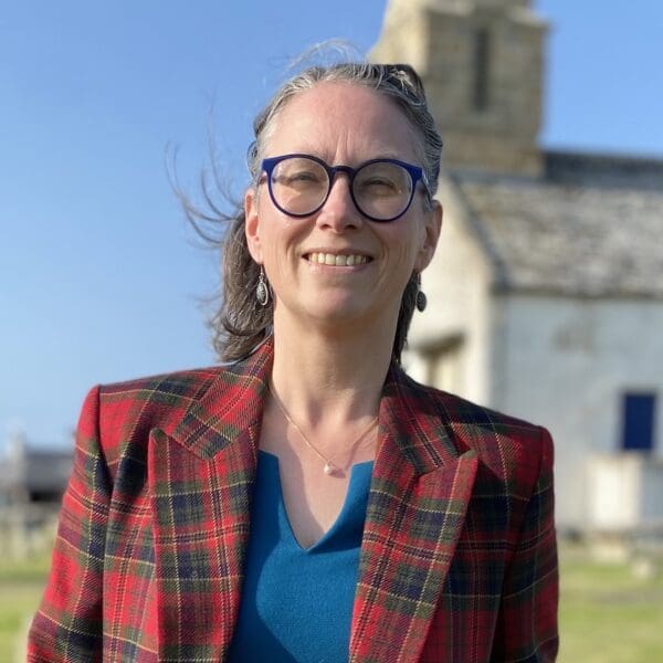 Head and shoulders image of female wearing red tartan jacket over blue top, standing in grass churchyard with white church in background