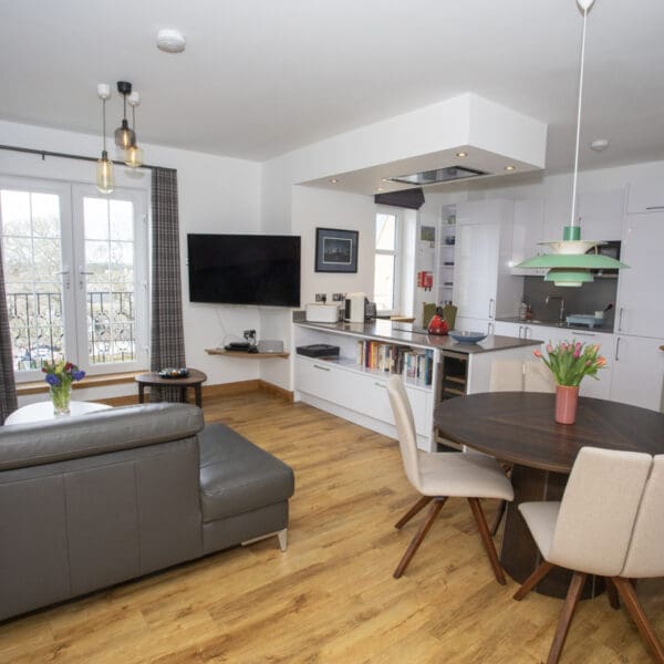 Open plan kitchen and living room with gloss white kitchen cabinets, oak floor boards, dark grey leather couch and circular dark wood dining table with cream chairs