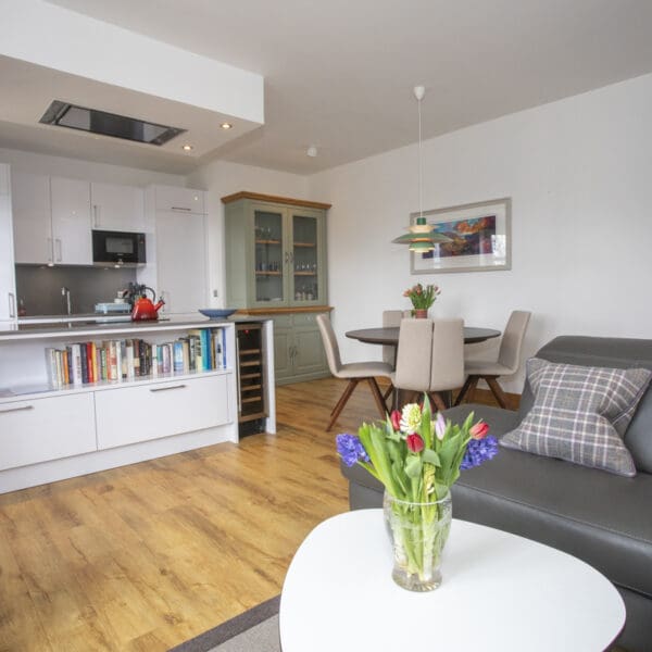 Open plan kitchen and living room with gloss white kitchen cabinets, oak floor boards, dark grey leather couch and circular dark wood dining table with cream chairs