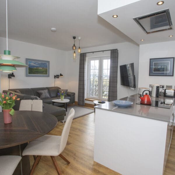 Open plan kitchen and living room with gloss white kitchen cabinets, oak floor boards, dark grey leather couch and circular dark wood dining table with cream chairs