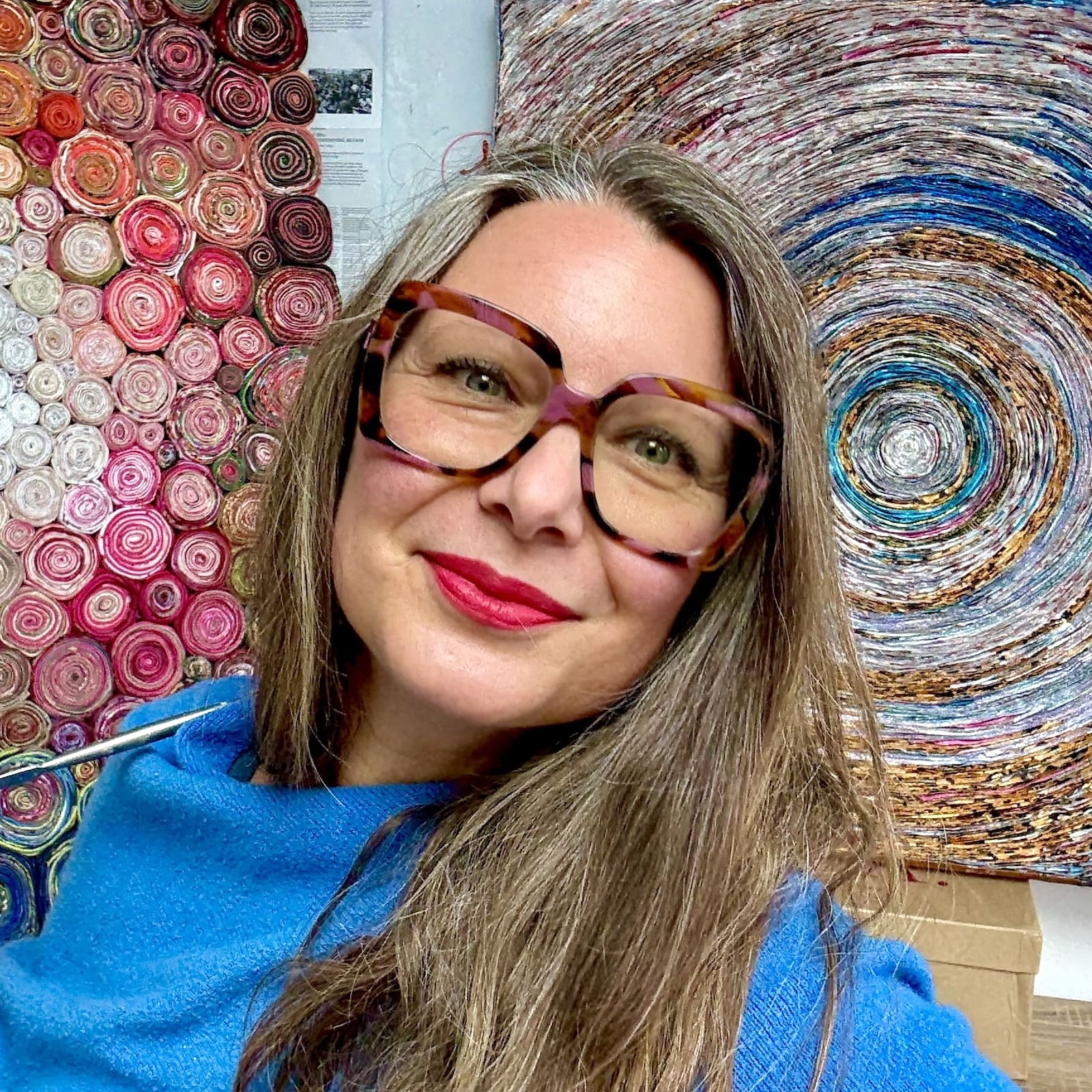 Image of female with long mid-brown hair wearing a bright blue top, large framed glasses and red lipstick, with multicoloured textile art in the background