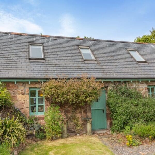 Exterior of stone cottage with slate roof. Green painted door and window frames. Climbing roses at door and green lawn in front