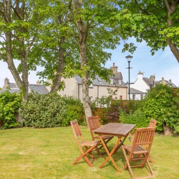 Wooden table and chairs in garden with grass lawn and mature trees, with neighbouring houses visible over hedge