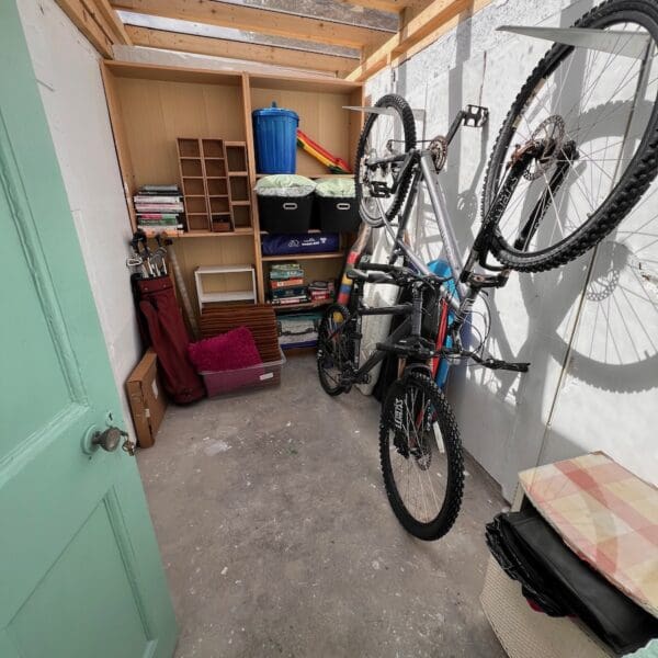 Storage shed with white walls and light green door, bikes hanging on wall and wooden storage shelves at rear