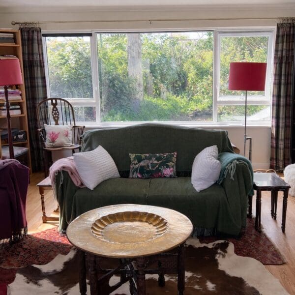 Living room with green couch set below large window looking out onto garden. Round wooden coffee table on white and black skin rug
