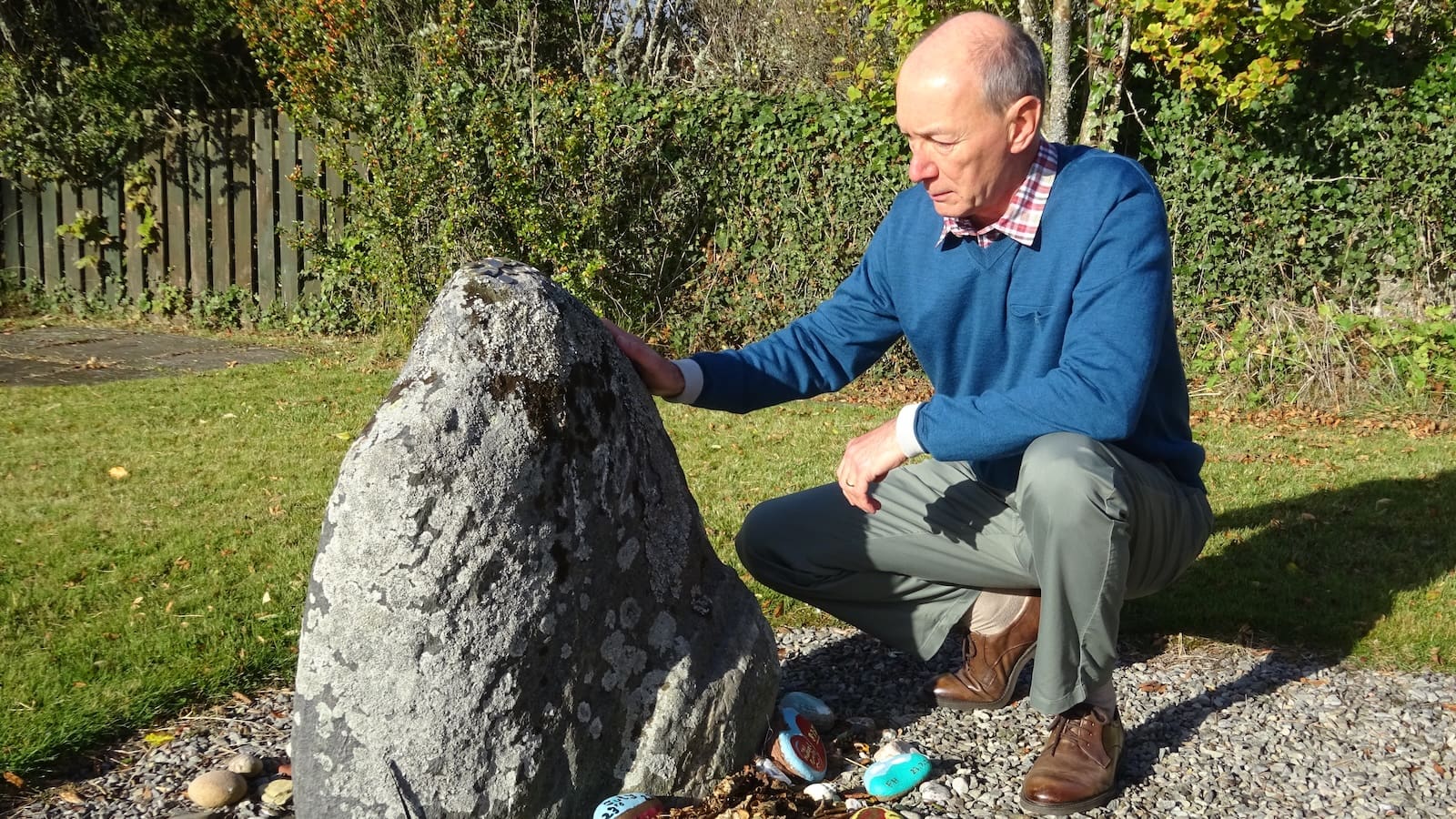 Man in blue jumper and grey trousers, crouching next to gravestone in garden with grass lawn and hedge in the background, witch's stone, Dornoch, Philip Paris author