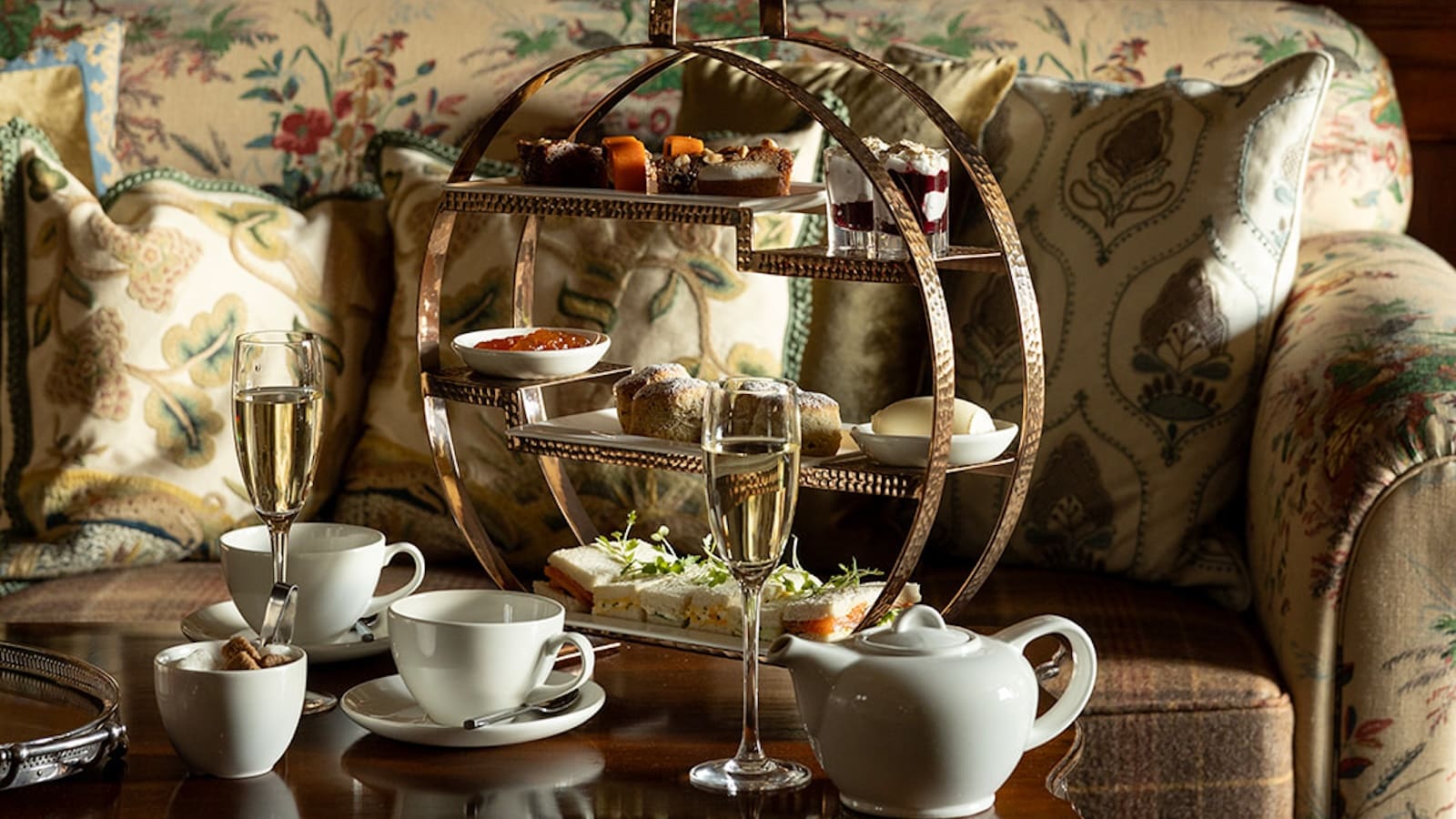 Afternoon tea of cakes and sandwiches on 3-tiered circular presentation tray, set on dark wood coffee table in front of floral sofa. White china tea set and Champagne flute