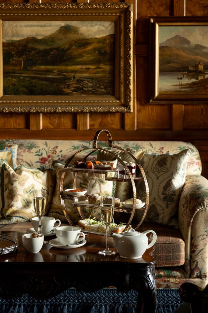 Afternoon tea of cakes and sandwiches on 3-tiered circular presentation tray, set on dark wood coffee table in front of floral sofa. White china tea set and Champagne flute