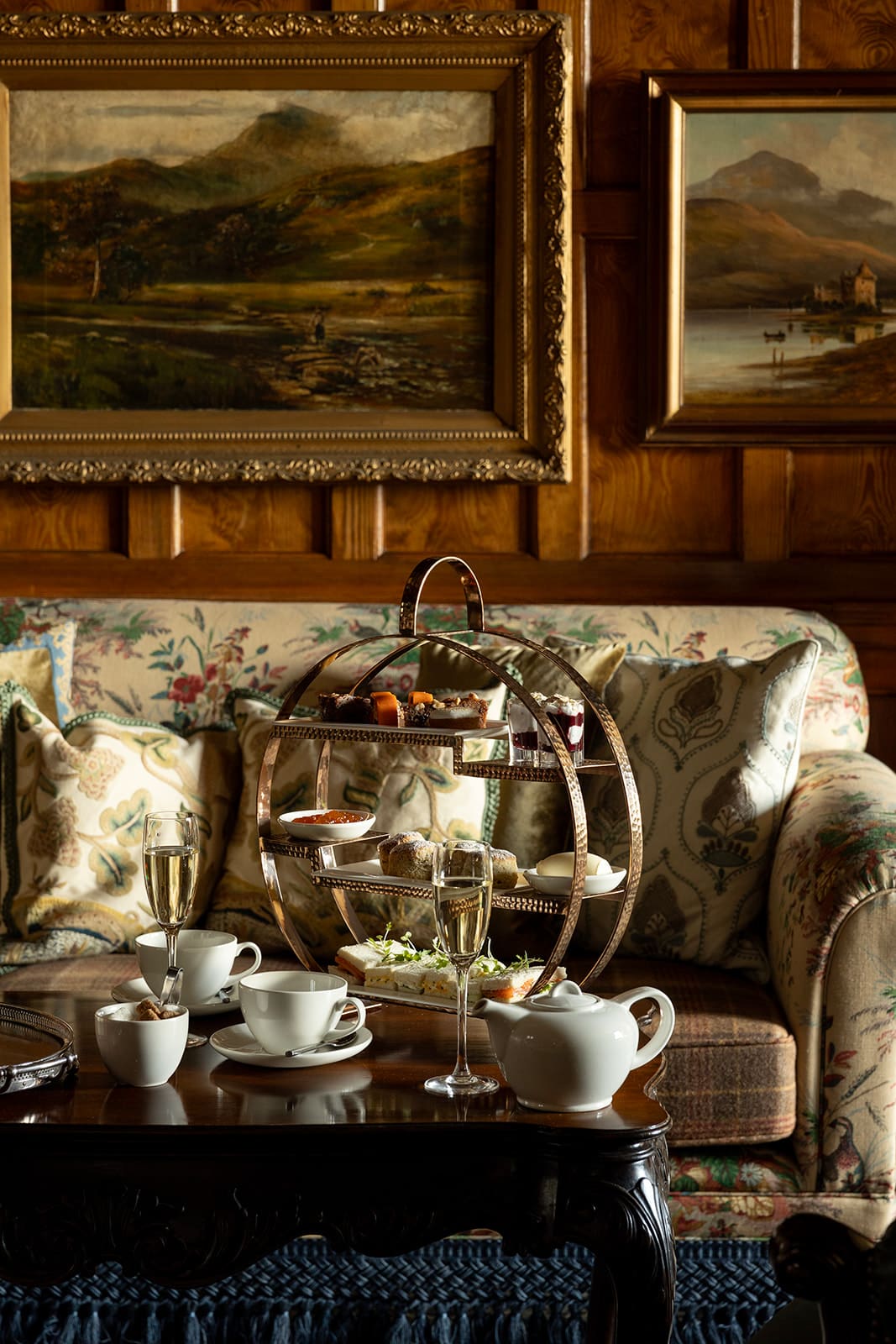 Afternoon tea of cakes and sandwiches on 3-tiered circular presentation tray, set on dark wood coffee table in front of floral sofa. White china tea set and Champagne flute