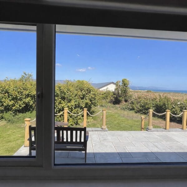 View out of large window over wooden decking out across garden to sea beyond