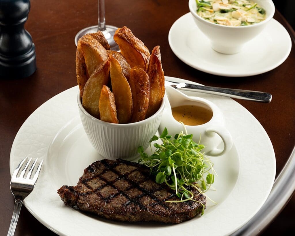 Seared stake on white plate with green salad, small jug of sauce and bowl of thick cut chips, set on dark wood tabletop alongside glass of red wine