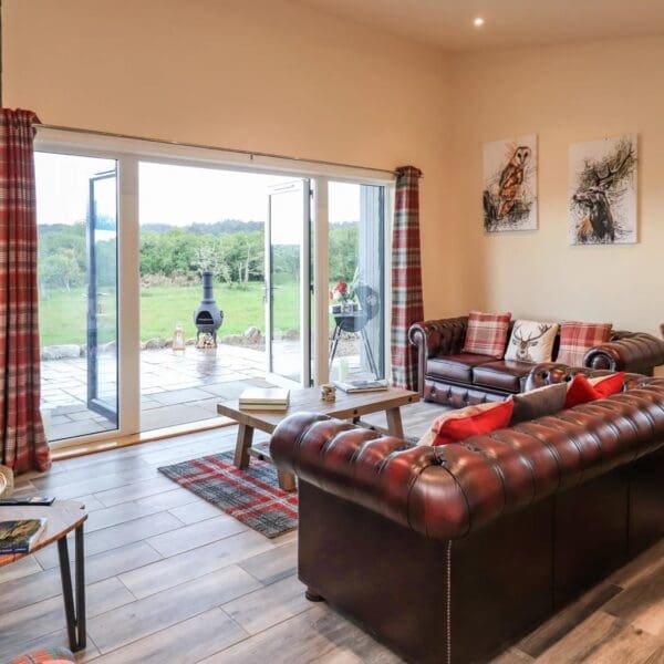 Living room with cream walls and red tartan curtains on large french doors overlooking patio area. Dark red leather Chesterfield style sofas with red accent cushions