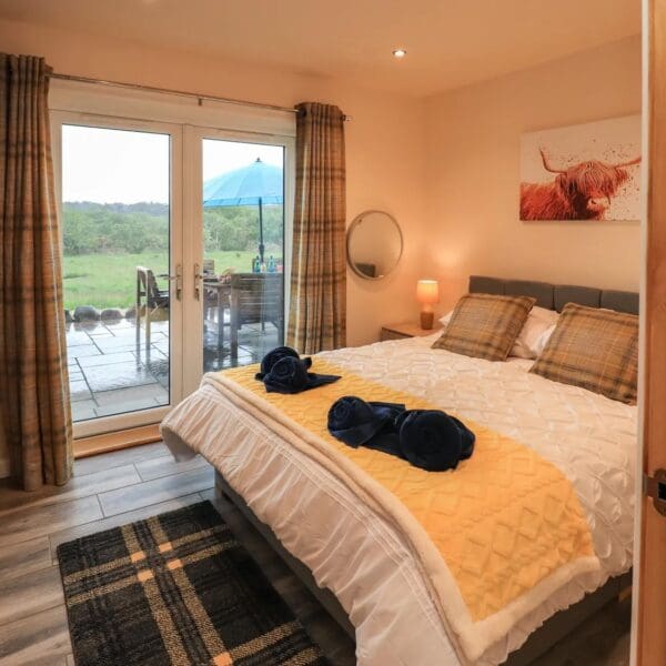 Double bedroom with cream walls and tartan curtains and rug. Patio doors overlooking grass field and woodland