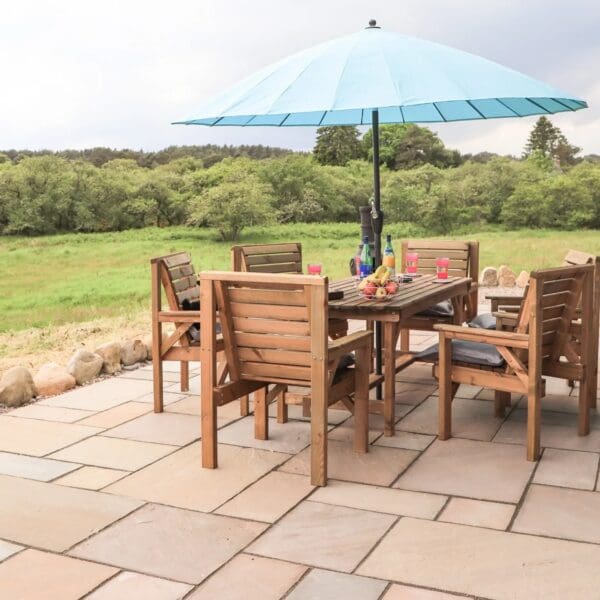 Paved patio area with wooden table and chairs and light blue parasol, overlooking grass field and woodland