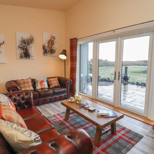 Living room with cream walls and red tartan curtains on large french doors overlooking patio area. Dark red leather Chesterfield style sofas with red accent cushions