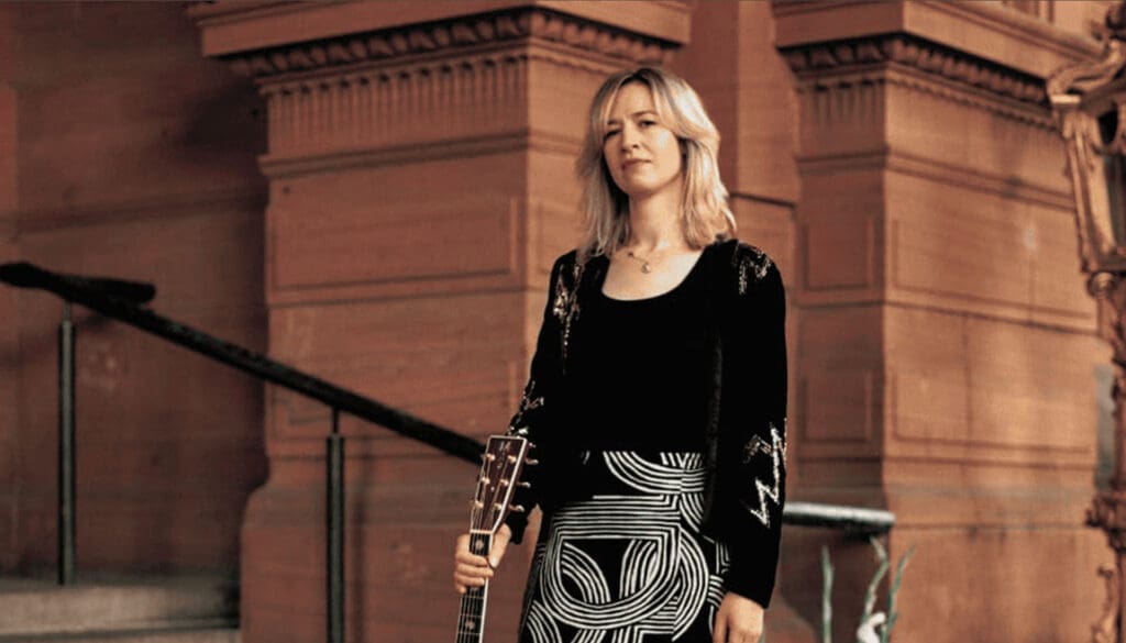 A photo of Jenn Butterworth standing in front of a flight of stone steps. She is holding a guitar.