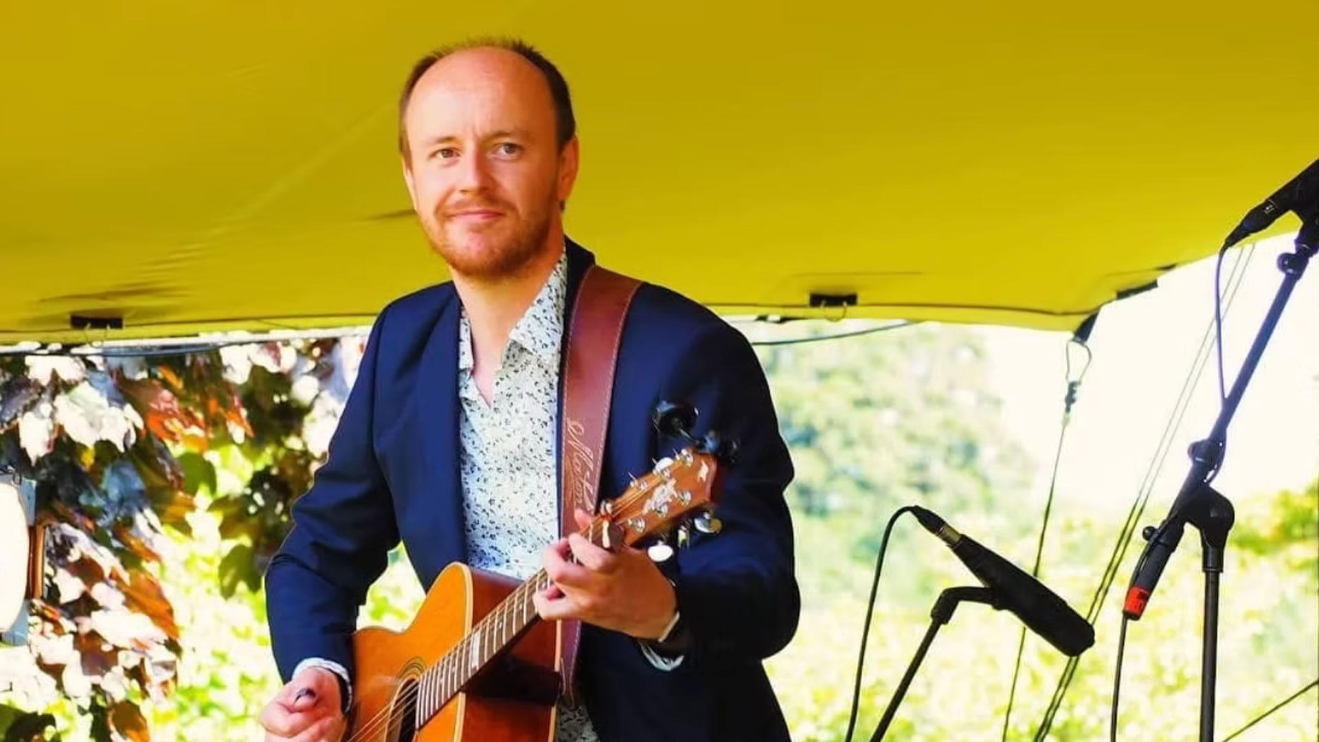 A photo of Liam Ross playing the guitar. In the background there is green trees
