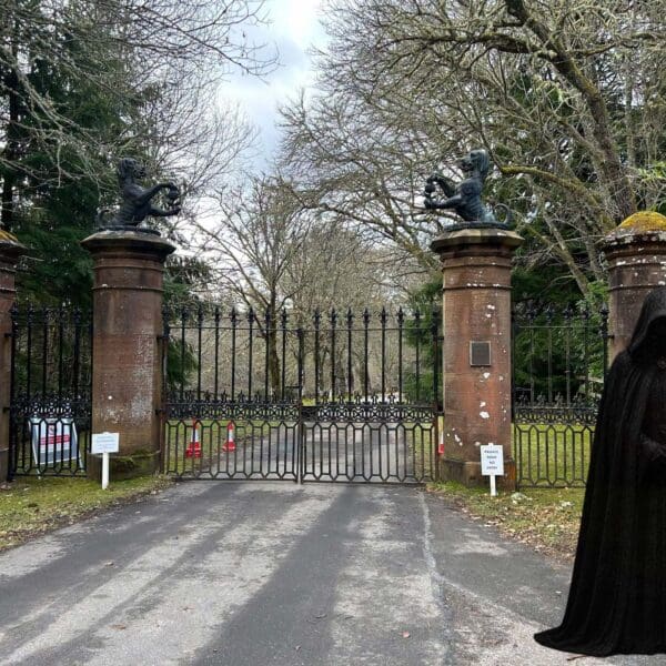 Photo of Ardoss Castle Gates showing hounds holding poppy seeds. To the right is a Traitor in a black cloak with their head bowed