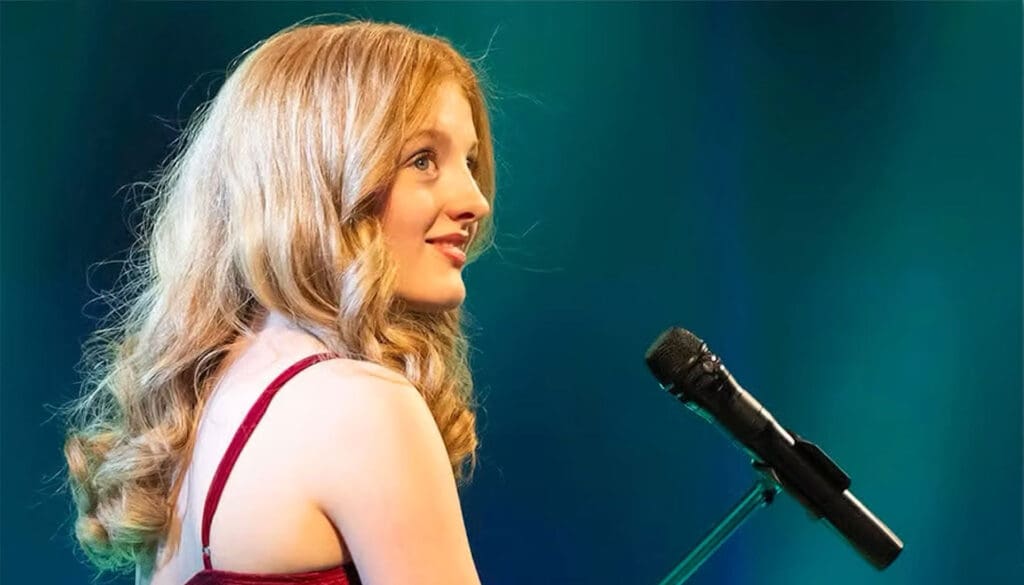 A photo of a white woman with blonde hair. She is sidewards on and in front of her is a microphone. She is wearing a strappy red dress and is smiling at the camera.